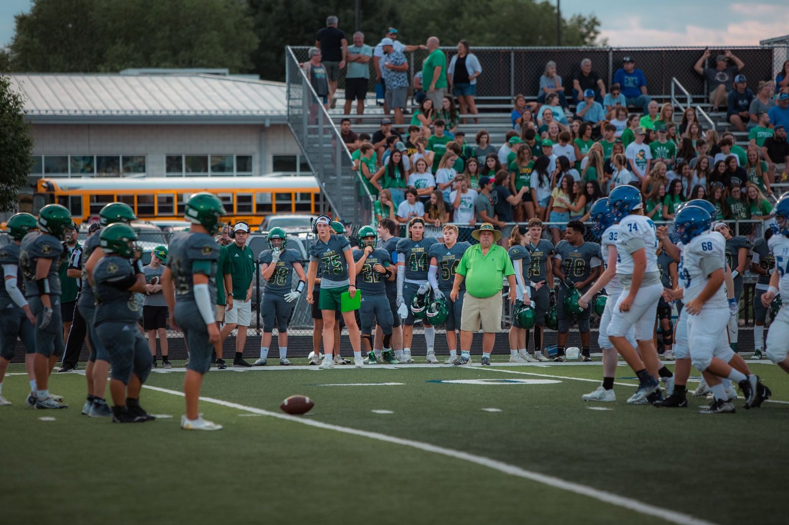 A packed high school football game — student section in school colors, yellow buses behind the bleachers